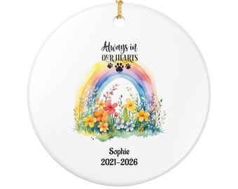May include: A round white ceramic ornament featuring a watercolour rainbow and colourful wildflowers. The text reads Always in OUR HEARTS with three black paw prints. Below the flowers, the name Sophie and the years 2021-2026 are printed in black.