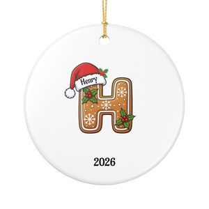 Puede incluir: Un adorno redondo de cerámica blanca con una letra H de pan de jengibre decorada con copos de nieve blancos, hojas de acebo verdes y bayas rojas. La letra lleva un gorro de Papá Noel rojo con el nombre Henry y el año 2026 debajo.