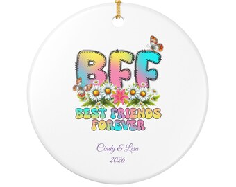 Puede incluir: Un adorno redondo de cerámica blanca con un cordón dorado para colgar. El diseño presenta el texto BFF en un degradado colorido, rodeado de margaritas y mariposas. Debajo, el texto dice BEST FRIENDS FOREVER, Cindy & Lisa y 2026.