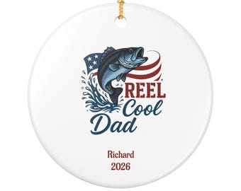 Puede incluir: Un adorno redondo de cerámica blanca con un pez azul saltando del agua, una bandera estadounidense estilizada y el texto REEL Cool Dad. Debajo del gráfico, el nombre Richard y el año 2026 están impresos en rojo. Incluye un cordón dorado.