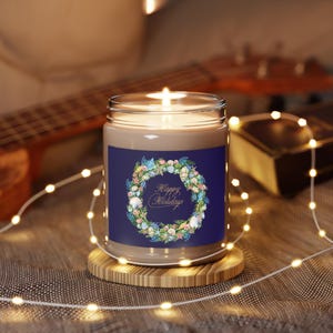 Peut inclure: Une bougie en pot de verre transparent avec une mèche allumée, posée sur un socle circulaire en bois. L'étiquette présente un fond bleu foncé avec une couronne de coquillages, d'étoiles de mer et de verdure, et le texte Happy Holidays en or.