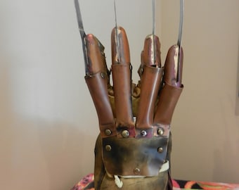 May include: A brown leather glove featuring four long, thin, silver-coloured metal blades attached to the fingers with copper-coloured metal plates and rivets. The glove is mounted on a rectangular base with a red and green striped design and a silver plaque reading Nightmare on Elm Street.