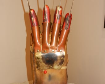 May include: A brown leather glove featuring four long, thin metal blades attached to the fingers. The glove has copper-coloured finger stalls, a metallic back plate secured with rivets, and a small red circular detail on the palm area.