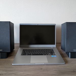 May include: A silver laptop with a black keyboard and a black screen is flanked by two black Philips speakers. The speakers are rectangular and sit on black bases. The laptop is open on a wooden surface.