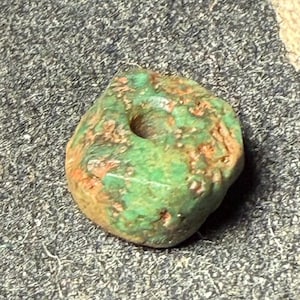 May include: A weathered, green stone bead with a central hole. The bead has a rough, textured surface with hints of orange and brown. The bead's shape is irregular, suggesting it is handmade or naturally formed.