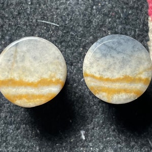 May include: Two round, polished stone plugs. The plugs have a gradient of colors, with a light blue and white top, transitioning to a yellow-brown band, and a white base. The plugs are likely for ear piercings.
