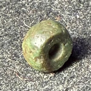 May include: A weathered, olive-green bead with a central hole. The bead has an irregular, rounded shape and a slightly textured surface. The color varies from light to dark green with some brown patches, suggesting age or natural variations. The bead is likely made of stone or a similar material.
