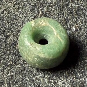 May include: A round, green jade bead with a hole in the center. The bead has a smooth, polished surface with variations in color. The jade is a light green color with some darker green and brown areas.