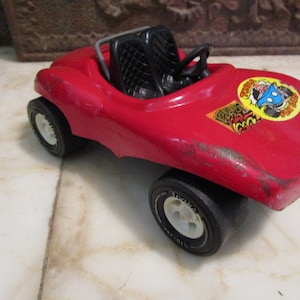 May include: Red toy car with black seats and white tires. The car has a sticker on the side that says "Zoom Fast Cars".