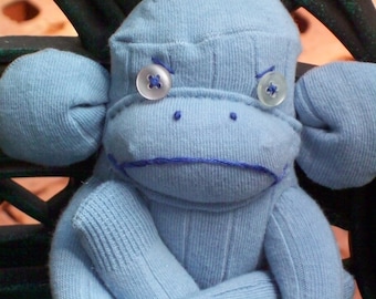 May include: A blue sock monkey with a sad expression. The monkey has two large buttons for eyes and a stitched-on mouth.
