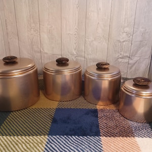 Może przedstawiać: Zestaw czterech miedzianych pojemników z pokrywkami. Pojemniki są wszystkie tego samego rozmiaru i kształtu, a mają błyszczące, metaliczne wykończenie. Pokrywki są wykonane z ciemniejszego materiału i mają lekko fakturowaną powierzchnię.