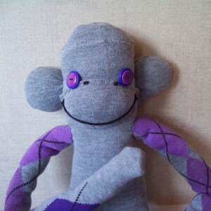 May include: A gray sock monkey with purple and gray argyle patterned arms and legs. The monkey has black eyes with purple buttons and a black stitched smile.