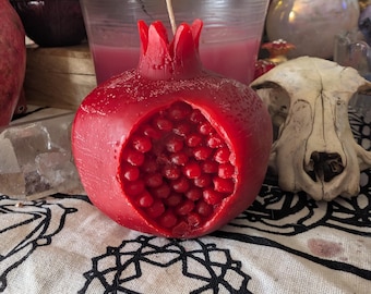 May include: A vibrant red pomegranate-shaped candle with a detailed interior, showcasing the seeds. The candle has a wick and sits on a patterned cloth. Other decorative items, including a skull and candles, are in the background.