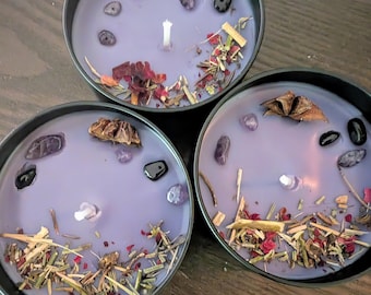 May include: Three round, black metal candles with lavender wax. Each candle is decorated with dried herbs, flowers, crystals, and a star anise. The candles have white wicks.