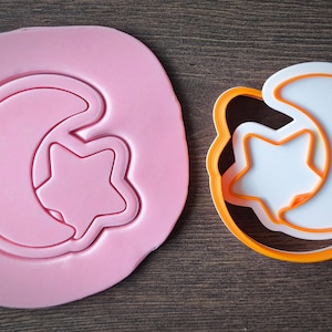 Op de afbeelding: Een roze fondant met een maan- en sterontwerp naast een witte en oranje koekjesvorm met hetzelfde ontwerp. De koekjesvorm is gemaakt van plastic en heeft een gedetailleerde omtrek van de maan en ster.