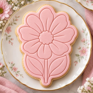 May include: A flower-shaped sugar biscuit decorated with smooth, light pink icing. The icing features embossed outlines of a central flower head with rounded petals and two leaves at the base, all centred on a white plate with floral patterns.