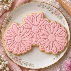Pode incluir: Um biscoito de açúcar grande em forma de três margaridas interligadas com cobertura lisa de cor rosa claro. O biscoito apresenta contornos de pétalas definidos em relevo e um centro circular para cada flor, sobre um prato de cerâmica branca.