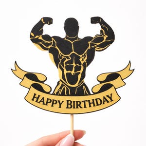 Pode incluir: Topper de bolo preto e dourado com a silhueta de um fisiculturista musculoso. A figura está a flexionar, com detalhes musculares dourados. Uma faixa dourada abaixo diz "HAPPY BIRTHDAY". O topper está preso a um palito de madeira.