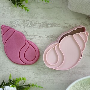 Può includere: Due stampini per biscotti a forma di conchiglia. Uno è di colore magenta scuro e l'altro è rosa pastello chiaro. Entrambi gli articoli presentano un design a conchiglia a spirale con bordi rialzati per tagliare e imprimere l'impasto.