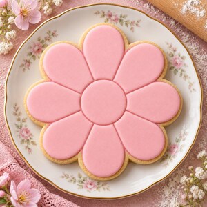 May include: A flower-shaped sugar biscuit with smooth, light pink icing covering the surface. The biscuit is centred on a white ceramic plate featuring a delicate floral pattern with small pink roses and green leaves along the scalloped gold rim.