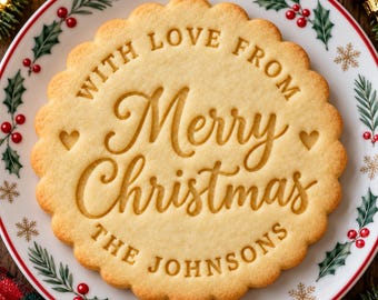 Op de afbeelding: Een rond koekje met geschulpte randen en de in reliëf aangebrachte tekst "WITH LOVE FROM Merry Christmas THE JOHNSONS" met twee kleine hartjes, in het midden van een wit bord versierd met groene hulstblaadjes, rode bessen en gouden sneeuwvlokken.