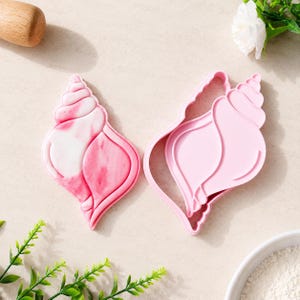 Può includere: Due stampini per biscotti a forma di conchiglia. Uno è di colore rosa chiaro uniforme, mentre l'altro presenta un motivo marmorizzato rosa e bianco. Entrambi gli strumenti hanno un bordo rialzato per tagliare e un design interno dettagliato.