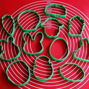 May include: A collection of green plastic cookie cutters in various Easter shapes, including a carrot, bunny, egg, and basket. The cutters are arranged on a red silicone baking mat with white measurement markings.