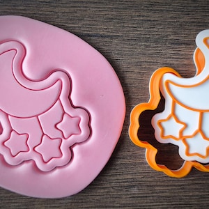 Op de afbeelding: Een roze siliconen mal en een oranje en witte koekjessteker in de vorm van een halve maan met hangende sterren. De mal is effen roze, terwijl de steker een witte binnenkant en een oranje omtrek heeft.
