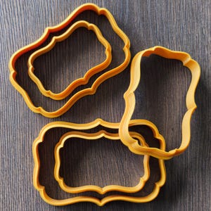 May include: A set of four orange cookie cutters, each featuring a rectangular shape with decorative, scalloped, and curved edges. The cutters are arranged in a cluster, showing varying sizes of the same ornate frame design with open centers.