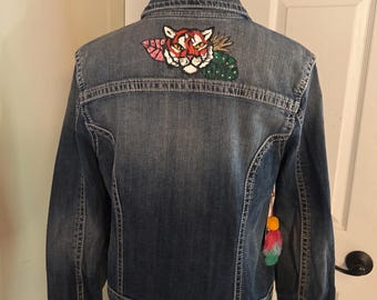 Könnte beinhalten: Eine blaue Jeansjacke mit einem bunten Tiger-Patch auf dem Rücken. Der Patch zeigt ein Tigergesicht mit roten, weißen und orangen Details, umgeben von grünen und rosa Akzenten. Ein dekorativer Quastenanhänger hängt an der Jacke.