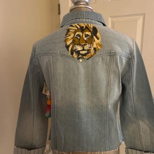 Puede incluir: Chaqueta vaquera azul claro con una cabeza de león bordada en la espalda. La chaqueta tiene un cuello clásico, cierre de botones y mangas enrolladas. La melena del león es una mezcla de dorado y marrón.