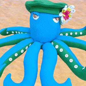 Könnte beinhalten: Ein blaues Plüsch-Oktopus-Spielzeug mit grünen Akzenten auf den Tentakeln und einem grünen Hut. Der Oktopus hat weiße Knöpfe und eine Blume. Der Hintergrund ist eine verschwommene sandfarbene Farbe.