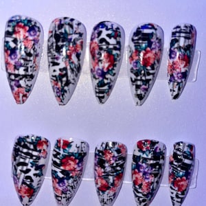 May include: Set of ten artificial nails with a floral design. The nails are white with a pattern of red, purple, and pink flowers, accented by black lines. The nails are arranged in two rows of five.