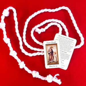 May include: A white knotted cord with a card featuring an image of Saint Joseph and a child, with the text "Pray for us." The card also includes text about the Cord of Saint Joseph on a red background.