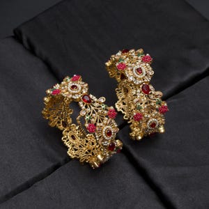 May include: Two gold bangles with detailed floral patterns. The bangles are decorated with red and green gemstones, and small white pearls. The jewellery is presented on a black satin surface.