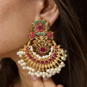 May include: Gold chandelier earrings with a floral design. The earrings feature red, green, and blue gemstones, and white pearls. The earrings are worn on an ear, showcasing the intricate details and craftsmanship.