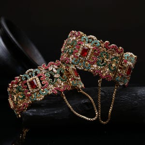 May include: Two ornate gold bangles with intricate designs. The bangles are adorned with red, green, and turquoise gemstones, along with small white pearls. A delicate gold chain connects the two bangles.