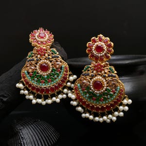 Peut inclure: Une paire de boucles d'oreilles dorées ornées. Chaque boucle d'oreille présente un dessus floral avec un grand centre de pierre rouge. Le corps principal a une forme de croissant avec des accents rouges, verts et turquoise, et de petites perles blanches.