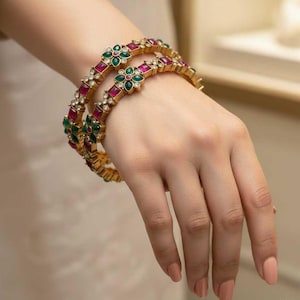 May include: Two gold bangles, each decorated with red, green, and clear gemstones. The bangles have a floral design with alternating square and flower-shaped settings. The jewellery is shown on a person's hand.
