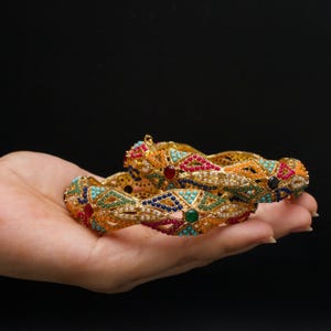 May include: Two gold bangles with detailed geometric patterns are held in a hand against a black backdrop. The bangles are decorated with red, blue, green, and turquoise gemstones, and small white pearls.