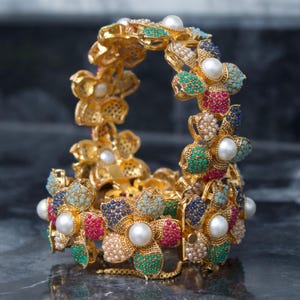 May include: A gold bracelet with a floral design. The bracelet is decorated with colourful gemstones in shades of red, green, blue, and white, and features large, round pearls. The bracelet is set against a dark background.