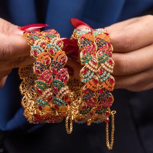May include: Two gold bangles with detailed leaf patterns. Each bangle is decorated with a variety of colourful gemstones, including red, blue, green, and orange, and small white pearls. The bangles are held against a dark blue background.