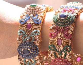 May include: Two gold bangles with elaborate designs, set with colourful gemstones. One bangle has blue and turquoise stones, the other green and pink. Both are decorated with pearls and intricate patterns.