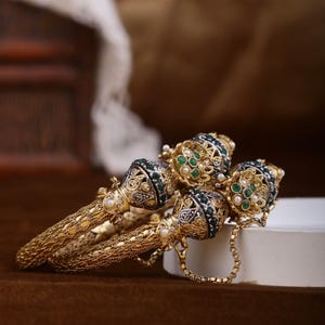 May include: Detailed gold bangles with an ornate design. The bangles have a textured surface and are embellished with green and white gemstones. The ends of the bangles feature a decorative, jewel-encrusted design.
