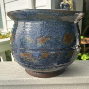 May include: A handcrafted ceramic planter with a rounded body and a wide, slightly flared rim. The planter features a dark blue glaze with brown and gold accents, and sits on a dark brown base. This decorative piece is suitable for indoor or outdoor use.