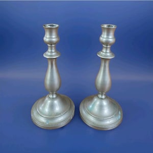 May include: A pair of matching silver-toned metal candlesticks featuring a classic turned design with a flared base, a slender central stem, and a wide, circular top cup for holding candles. The metal surface has a smooth, matte finish.