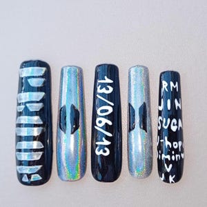 May include: Five long, artificial nails with various designs. One nail has the word "LOVE" in white, another has a holographic design with a black logo, and a third displays the date "13/06/13". The last nail has the names "RM, JIN, SUGA, J-Hope, Jimin, V, JK".