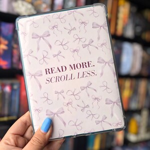 May include: A clear tablet case with a light pink bow pattern and the text "READ MORE. SCROLL LESS." The case is held in front of a bookshelf. The bows are in various sizes and orientations.