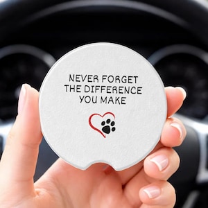 May include: A round white car cup holder coaster featuring a small notch at the bottom. The text reads NEVER FORGET THE DIFFERENCE YOU MAKE in black capital letters, accompanied by a red heart outline and a black paw print graphic in the center.