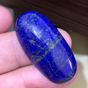 Puede incluir: Una gema de lapislázuli pulida, de forma ovalada. La piedra azul oscuro tiene motas y vetas doradas en su superficie. La gema es lisa y refleja la luz, mostrando su belleza natural. Se utiliza a menudo en joyería y artículos decorativos.